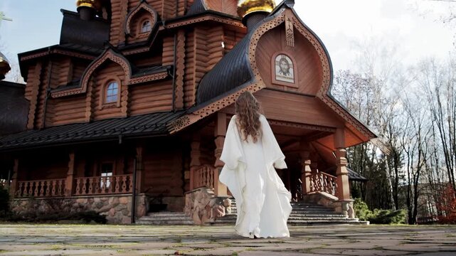 Caucasian woman with long curly hair, wearing flowing white dress, walks on stone path towards magnificent wooden church. Architecture features ornate carvings, multiple golden domes, and stone founda