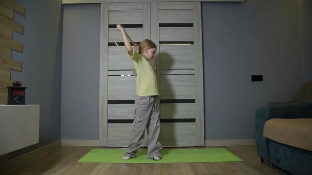 Young girl performs warm-up. Wooden stick behind shoulders. Torso rotation for flexibility. Green yoga mat, wood floor. Home interior for child fitness.