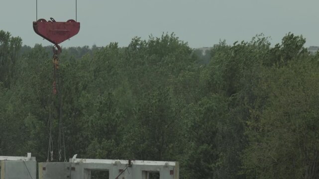 Close up view of powerful red industrial crane hook slowly lowering heavy pre-fabricated concrete wall panel into position for new building construction with dense green forest trees in background