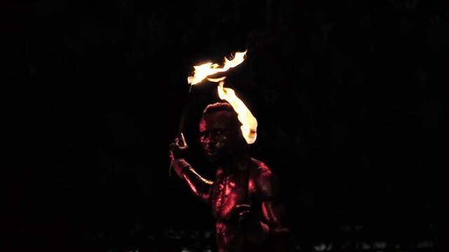 Dynamic fire artist performance in low light, manipulating multiple lit torches against dark background. Performer demonstrates mastery of fire techniques for engaging spectacle. Body reflects intense