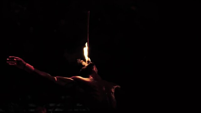 Fire eating. Black fakir performs an enthralling night show, demonstrating mastery over fire. Person holds a flaming torch, exhales fire, and balances a lit stick on face. Dark background accentuates 