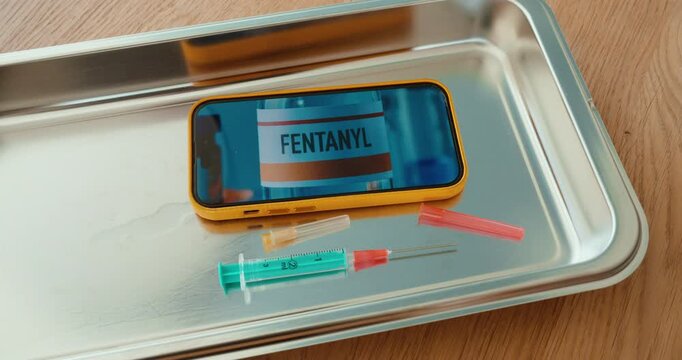 Hand placing a syringe next to a smartphone displaying a fentanyl label on a metal tray. Concept for opioid crisis, drug abuse, overdose risk, and public health awareness