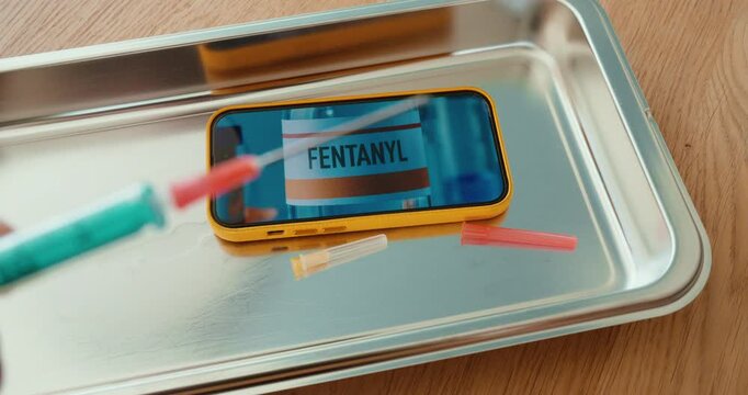 Close-up of syringes around a smartphone showing a fentanyl label on a metal tray. Concept for opioid crisis, drug abuse, overdose risk, and public health awareness.