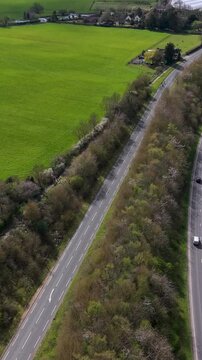 A414 road vertical drone flyover showing a forward approach to a T-junction with traffic, fields and woodland verges. Panshanger quarry visible to the right in Hertfordshire, England.