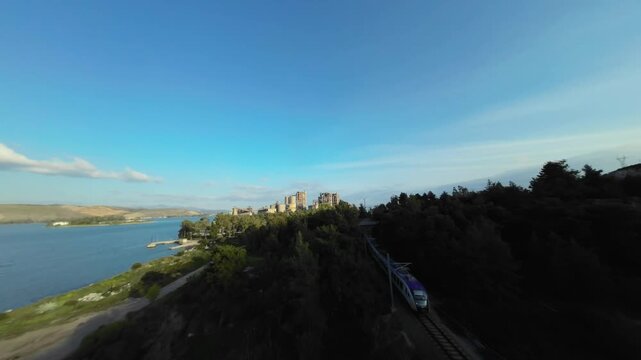 High-speed FPV drone dive through the abandoned Chalkida cement factory. The footage tracks through industrial ruins and trees to reveal the suburban train crossing beneath the High Bridge.