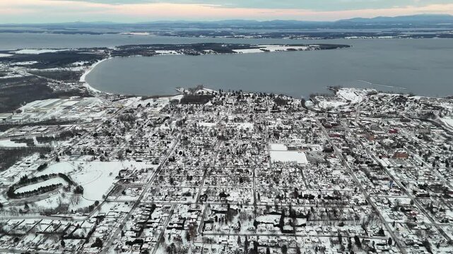 High altitude aerial view flying over the snow covered city of plattsburgh in upstate new york, showing the city grid, cumberland bay, and lake champlain with the green mountains in the distance
