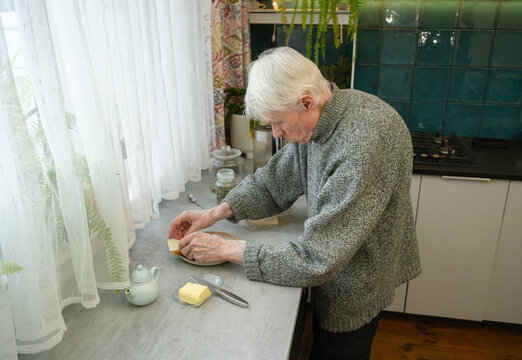 An elderly person with a curved spine makes sandwiches at kitchen counter. The image reflects physical effects of aging and importance of self-reliance. Grandpa engaged in task.