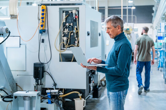 Senior engineer using laptop in a production hall with a young technician in the background