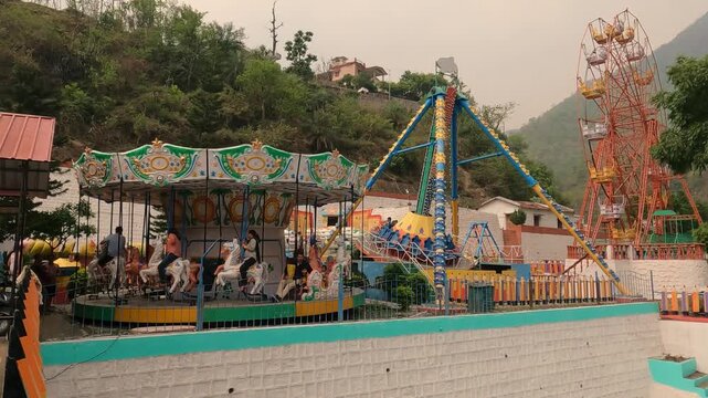 Colorful amusement park with carousel, swing ride and ferris wheel set against a scenic hillside. People enjoying leisure activities in a small hill station fairground.