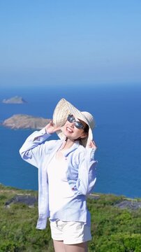 Asian woman enjoying summer vacation at the beach, wearing sun hat and sunglasses, smiling with blue ocean background