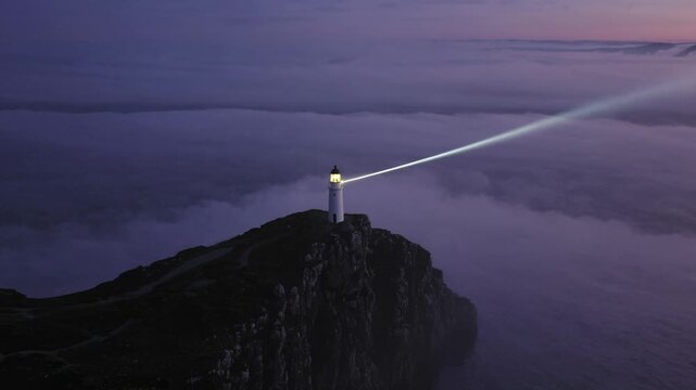 Lighthouse on rocky cliff emits bright beam of light across foggy ocean at twilight, illuminating the surrounding mist and creating a dramatic coastal scene