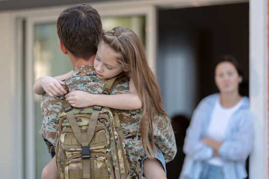 Emotional moment of soldier father holding daughter with backpack outside home, military deployment or return concept, family separation, love and protection, parenting bond, farewell scene.