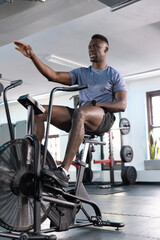 African American man pedaling and gesturing on fan air bike at gym, wearing shorts and watch © wavebreak3