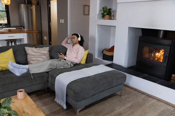 Obraz premium Non-binary adult reclining on dark gray sofa at home, holding smartphone, listening with headphones
