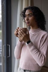 Nonbinary adult standing near window holding mug, wearing pink sweater, smartwatch, gazing at rain © wavebreak3