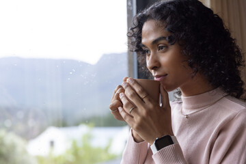 Indian nonbinary holding beige mug, wearing turtleneck and rings, gazing out wet window, copy space © wavebreak3