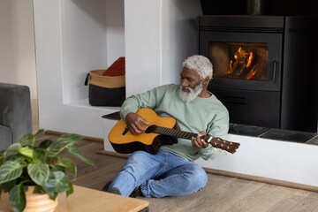 Senior African American man sitting on wooden floor playing acoustic guitar by wood-burning stove © wavebreak3