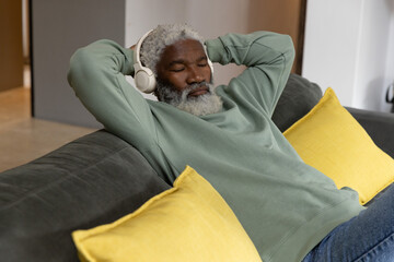 Senior African American man reclining on sofa at home, listening with headphones, hands behind head © wavebreak3
