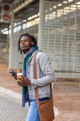 African American man waiting at bus stop wearing denim jacket knit scarf holding phone, coffee © wavebreak3