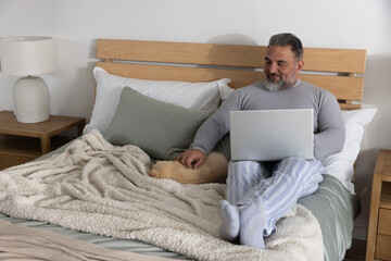 Mature man sitting propped against wooden slatted headboard on bed, petting dog and using laptop © wavebreak3