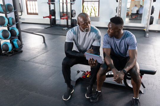 African American senior and adult men in athletic gear reviewing plan on gym bench with tablet