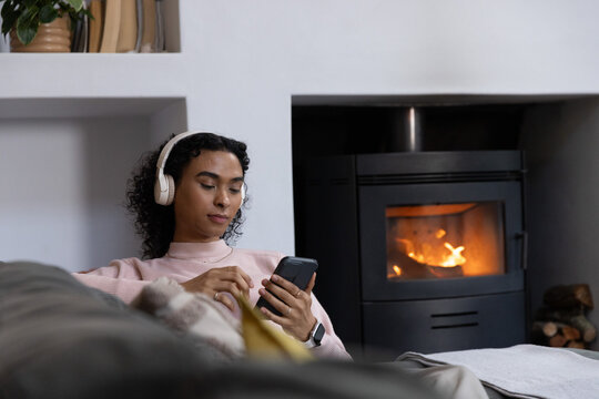 Nonbinary adult reclining on sofa at home wearing headphones and smartwatch holding phone by stove