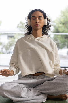 Non-binary adult meditating crosslegged on green mat wearing cream hoodie gray sweats headphones