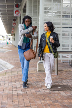Diverse friends walking at transit stop platform, one checking smartphone other holding coffee cup