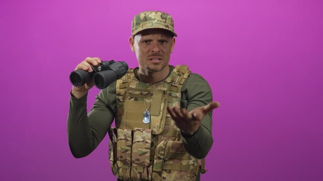 Man soldier in camouflage vest and cap pointing finger while holding binoculars in studio with purple backdrop; vigilance duty.