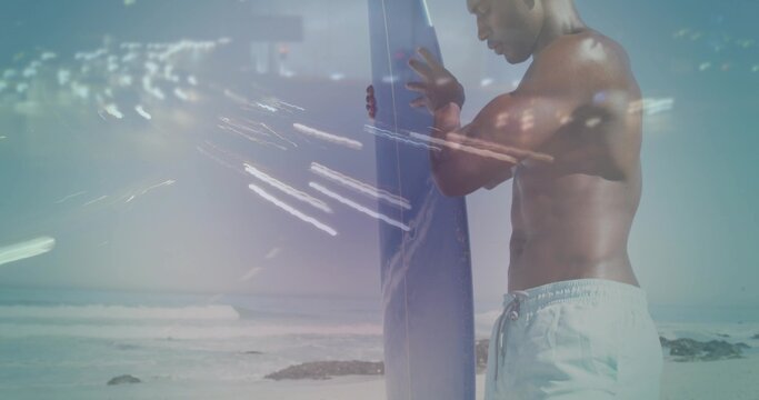 Standing shirtless surfer holding board on beach in light trunks, waves, rocks, flare, copy space