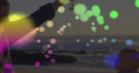 Fototapeta premium Caregiver and infant interacting as caregiver lifting arm, releasing sand with bokeh at dusk beach