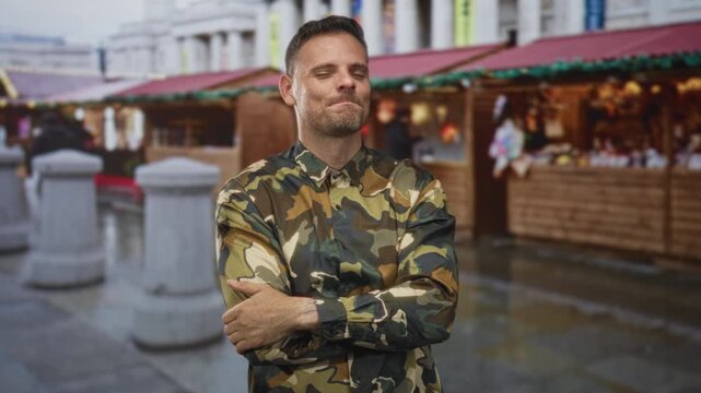 Man in camouflage shirt with arms crossed and a slight smile on street near market stalls; confidence duty.