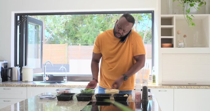 African-American man on call packing black meal boxes rice, veg at island checking phone smiling