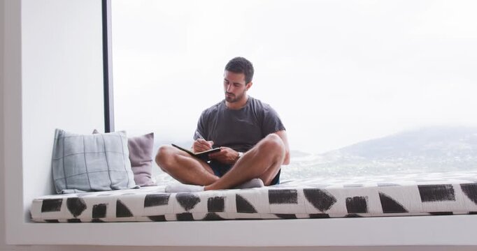 Man settling on built-in window bench and writing with pen in notebook, glancing at hills