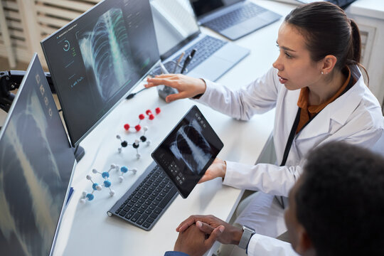 Young adult Caucasian female doctor reviewing chest x rays on tablet and monitor while consulting colleague. Supporting medical diagnosis, radiology workflow, healthcare technology marketing