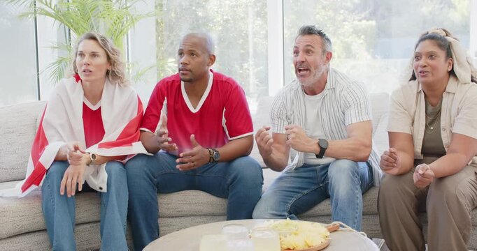 Diverse friends leaning forward, reacting to late game goal on sofa with flag, smartphone, snacks