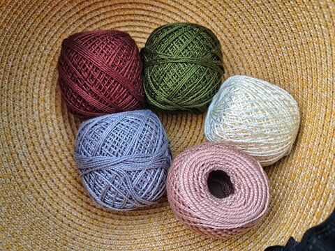 Five spools of vibrant mercerized cotton yarn in pink, blue, green, and white, arranged inside a woven straw basket for craft projects.