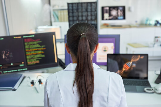 Rear view of young adult woman scientist analyzing genetic data across multiple screens in laboratory, supporting genomics research and biotech innovation marketing, wide shot