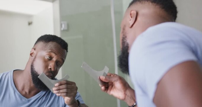 African man holding curved beard stencil, aligning and trimming with razor at mirror for neat edge