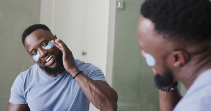 Man wearing T-shirt applying light-blue under-eye patches at bathroom mirror, checking reflection