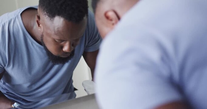 Man in blue shirt leaning at sink rinsing mouth, spitting, puffing cheeks, checking face in mirror