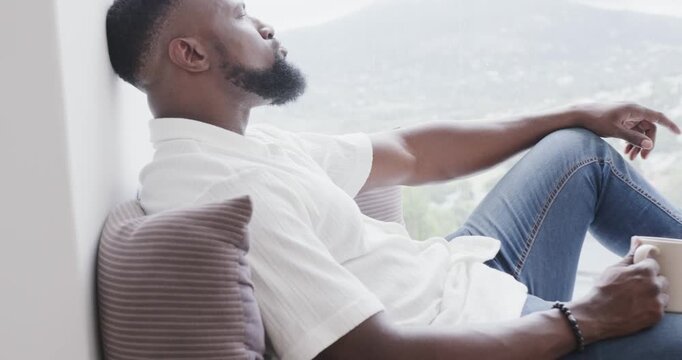Gazing from window bench thinking, African American man reaching for mug, showing beaded bracelet