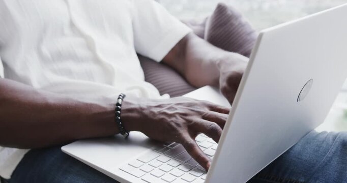 African American man typing on white laptop by large window, wearing white headphones and bracelet