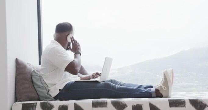 African American adult male typing on laptop at window bench, hearing cue, adjusting headphone
