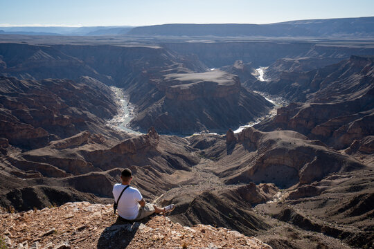 View of a man sitting on the edge of a precipice gazing upon the Fish River Canyon's vast, arid landscape under a clear sky, Keetmanshoop, Karas Region, Namibia.
