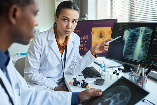 Portrait of Asian female doctor discussing lung scan with Black male colleague, reviewing diagnostic imaging on monitors. Ideal for healthcare teamwork, radiology, diagnosis marketing