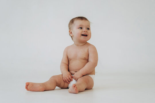 Baby sitting on a white background, wearing only a diaper, smiling and looking to the side, showcasing soft skin and chubby limbs in a bright, minimalistic setting