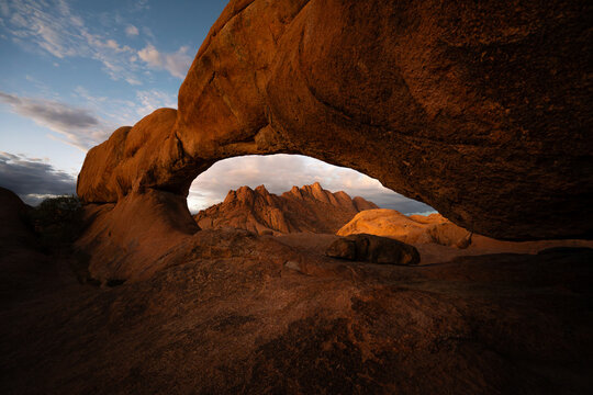 View of the rugged, sun-kissed archway framing distant peaks under a serene sky, capturing the raw beauty of the arid landscape, Spitzkoppe, Erongo Region, Namibia.