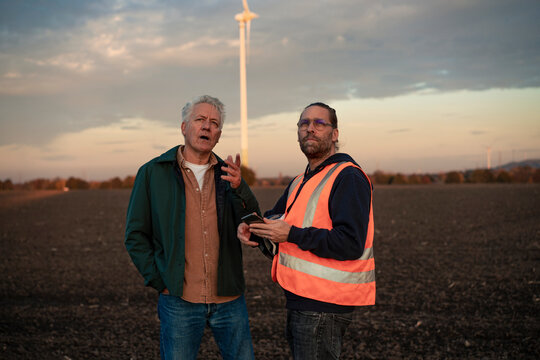Two industry workers discussing energy economy outdoors near a wind turbine at sunset