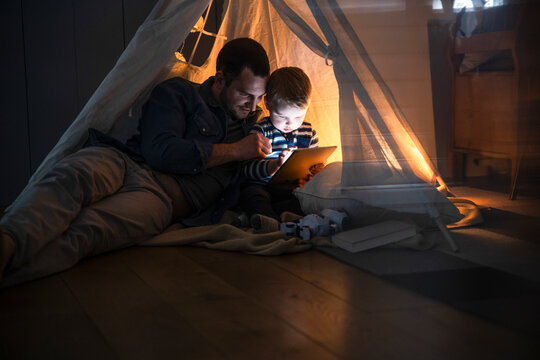 Father and son reading on tablet in cozy tent at home at night